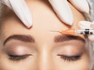 a woman with her eyes closed and a specialist preparing to inject Botox into her forehead 